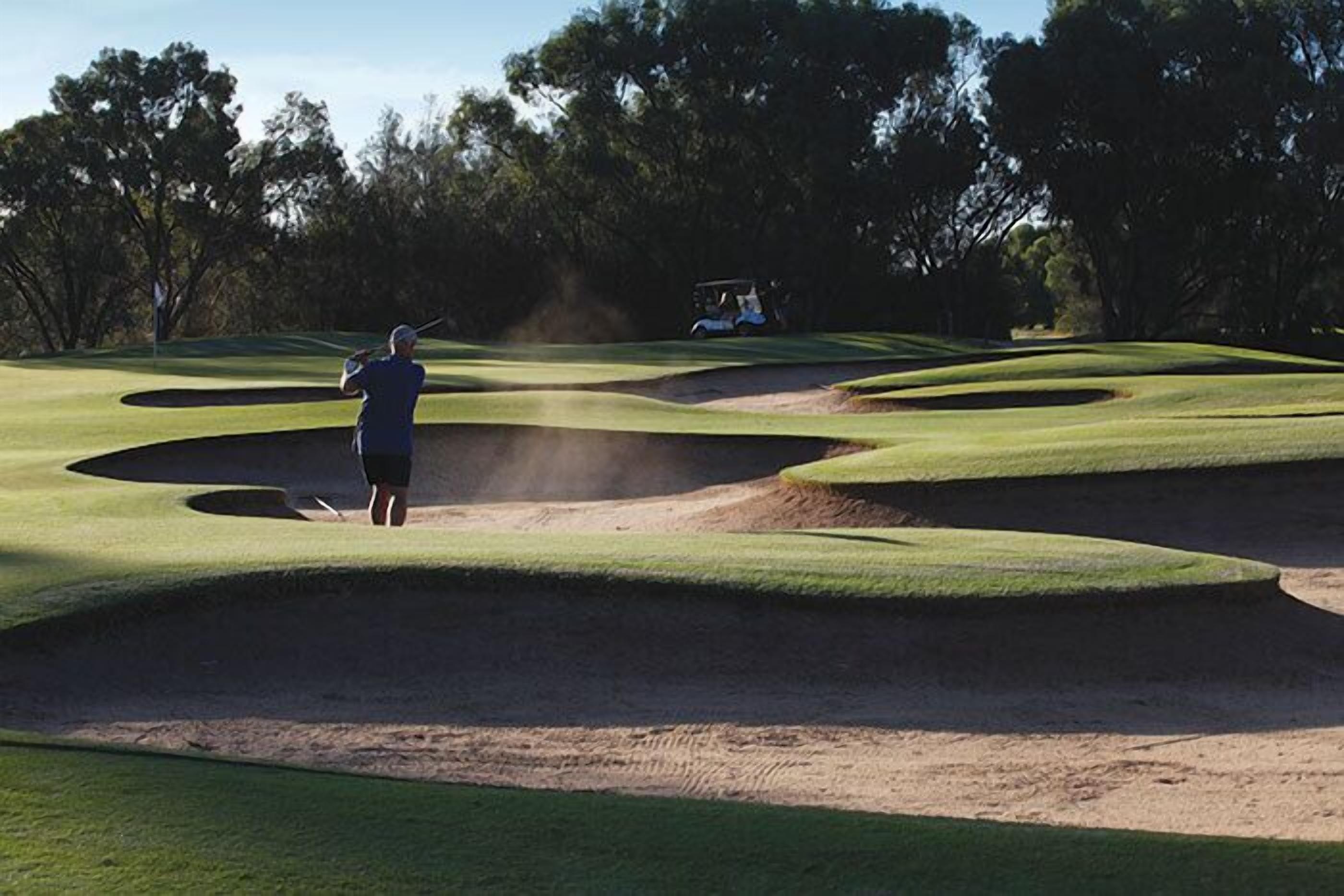 Campo de golf con un golfista a punto de golpear, un carrito de golf al fondo y cielo despejado.