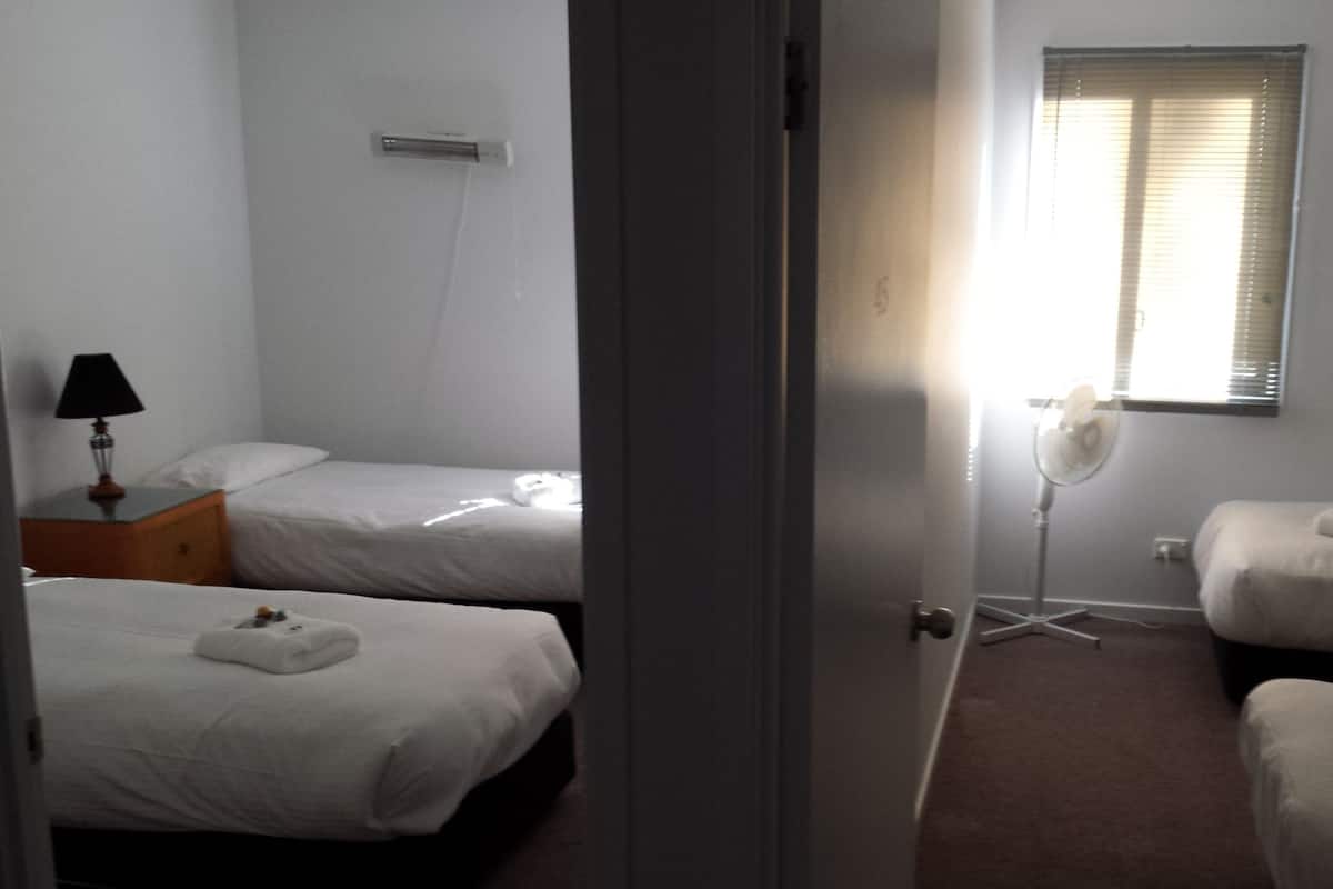 A hotel room with two beds, a bedside table with a lamp, a standing fan, and a window with blinds.