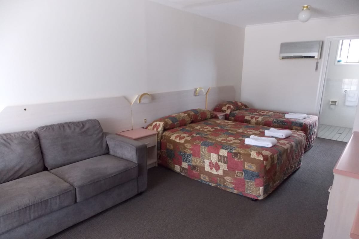 Una habitación de hotel con un sofá gris, dos camas individuales con colchas estampadas, una mesita de noche con lámpara y un ventilador de techo.