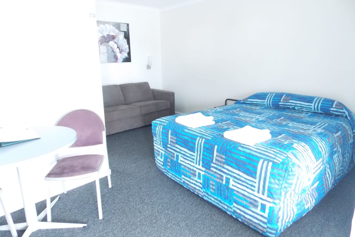 Una habitación con cama, una silla, una mesita, un sofá y un cuadro en la pared.
