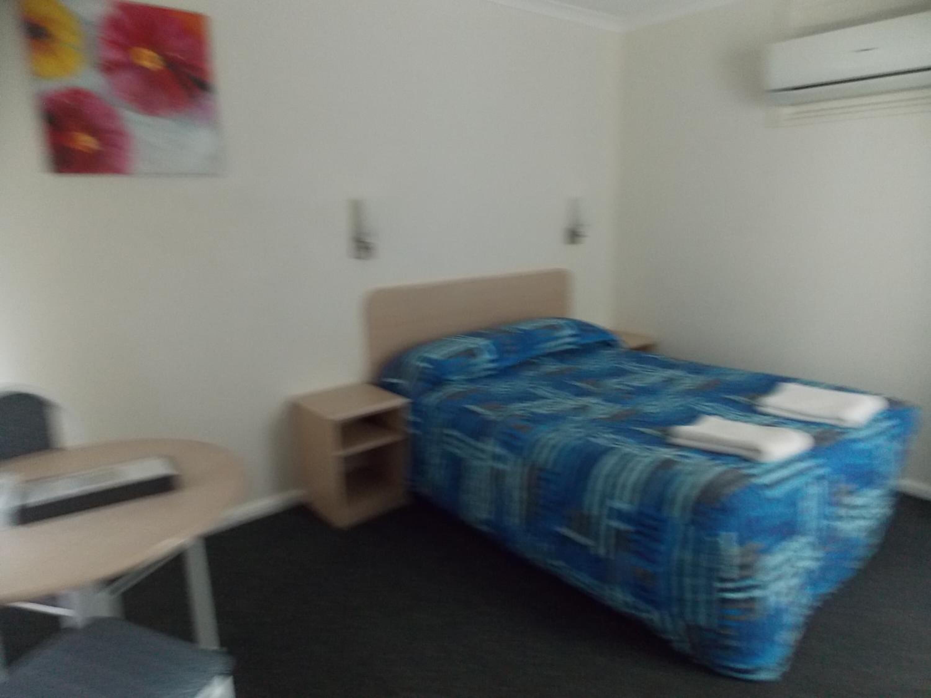 Ein kleines, schlichtes Zimmer mit einem Bett, einem Schreibtisch und einem Stuhl. Es gibt einen Nachttisch und eine Wand-Klimaanlage.