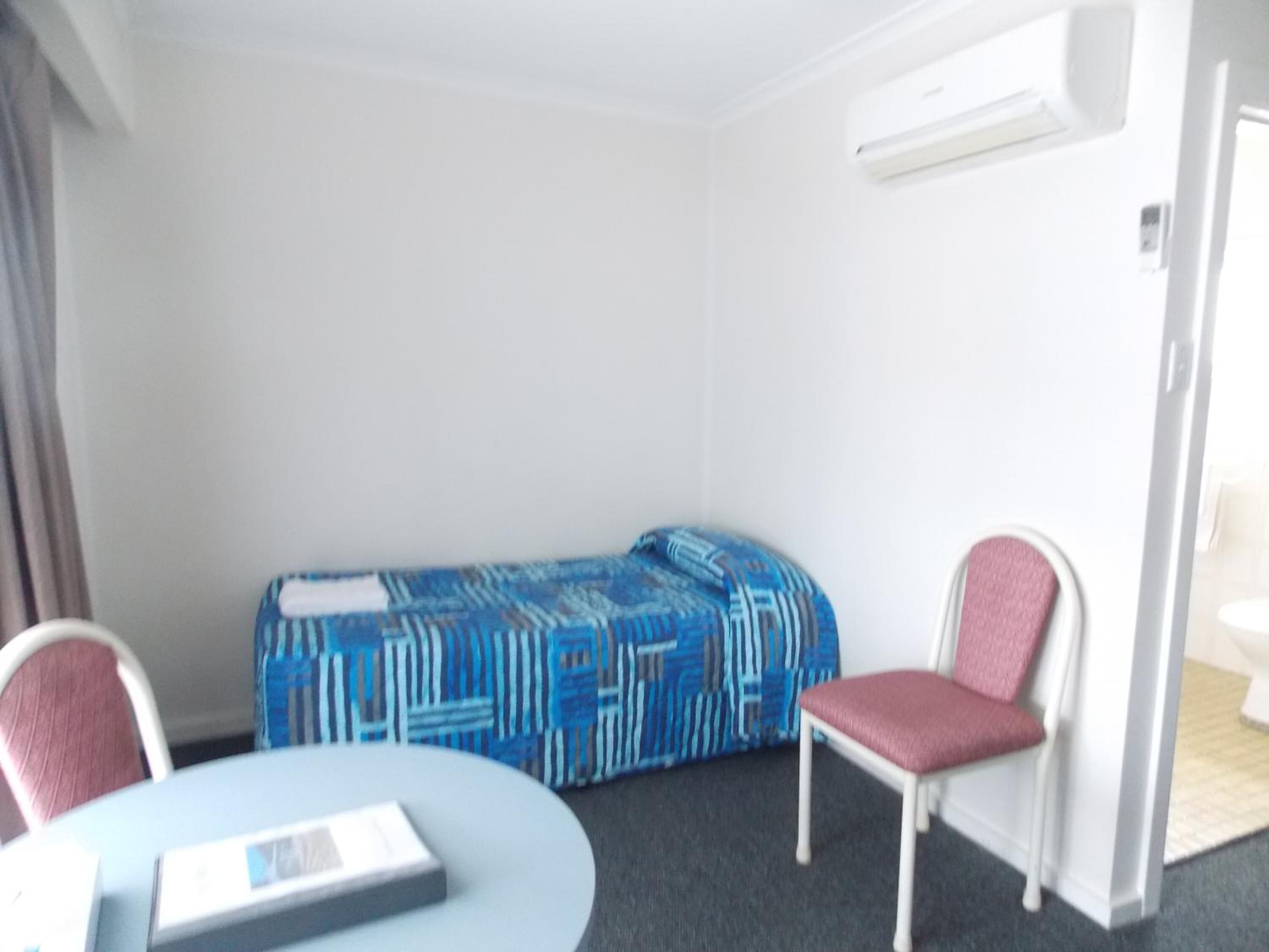 Una habitación de hotel pequeña y sencilla, con una cama, una mesa redonda, dos sillas y una mesita de noche con un libro.