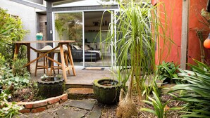 Un patio moderno al aire libre con una mesa y sillas de madera, un pequeño jardín con plantas y un camino de piedra.