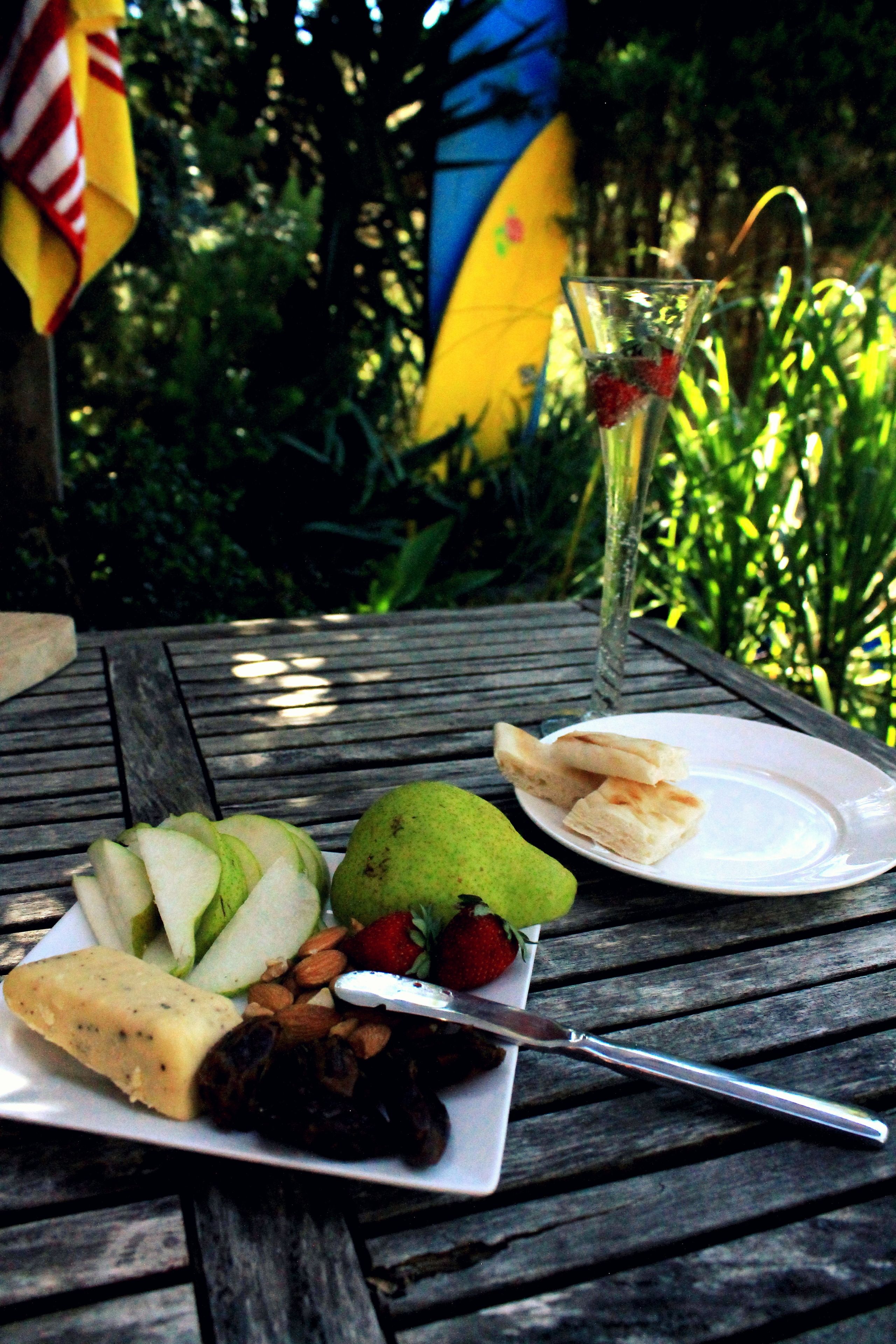 A meal on a wooden table outdoors with a glass of drink, fruits, and cheese.