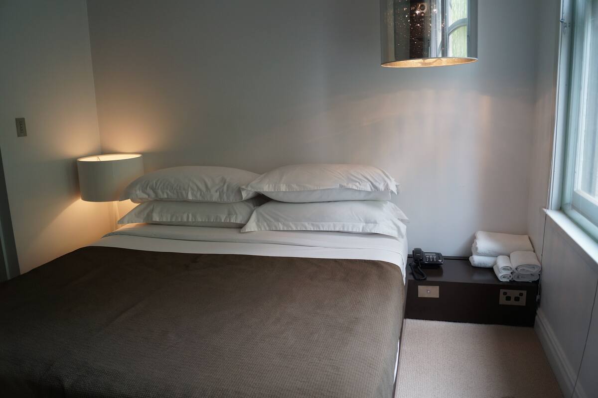A neatly made bed with white linens, a bedside lamp, a phone, and towels on a small table beside the bed.