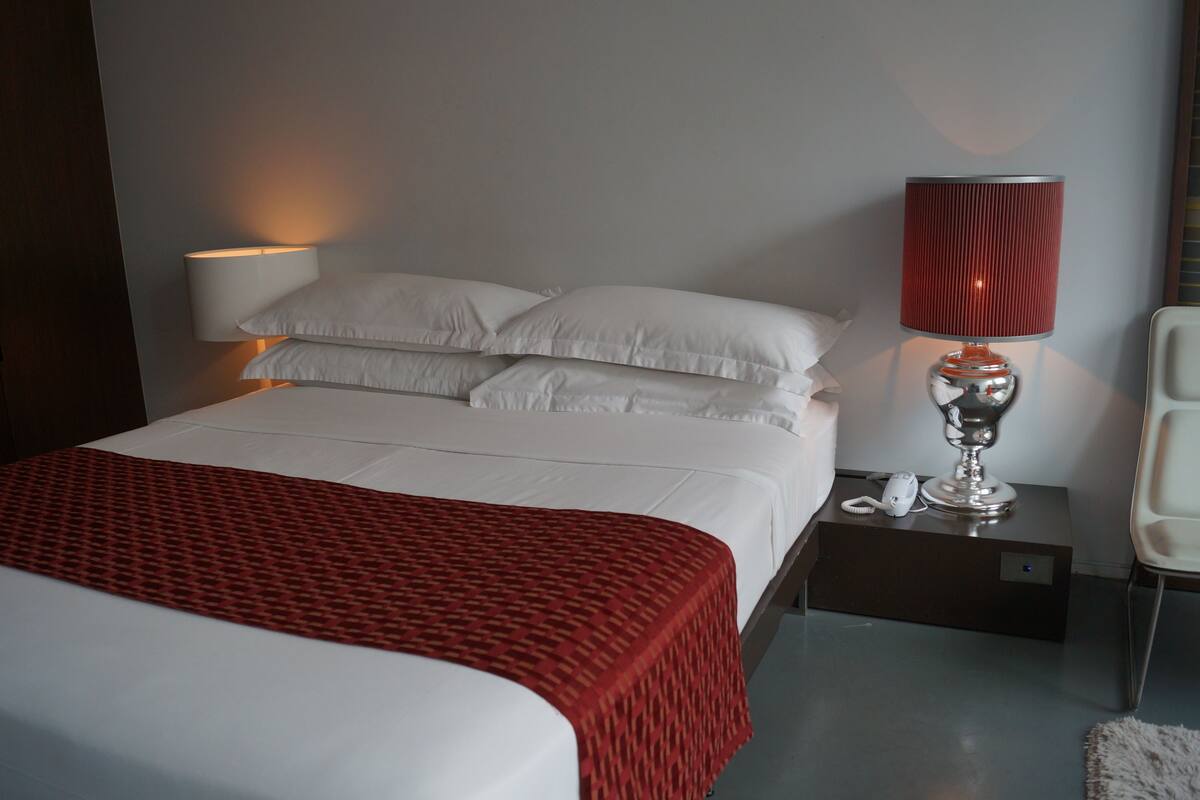 A neatly made bed with white linens and a red patterned bedspread, a bedside lamp, a white chair, and a small table with a phone and a charger.