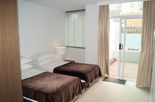 Quarto de hotel com duas camas, uma janela com persianas, uma porta com puxador e vista para um edifício com a inscrição 