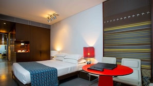 A modern hotel room with a neatly made bed, a red table with a remote control, and a wall with a striped pattern. - Valentine On George (Haymarket)