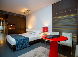 Quarto de hotel moderno com uma cama bem arrumada, uma mesa vermelha com um controle remoto e uma parede com padrão listrado.