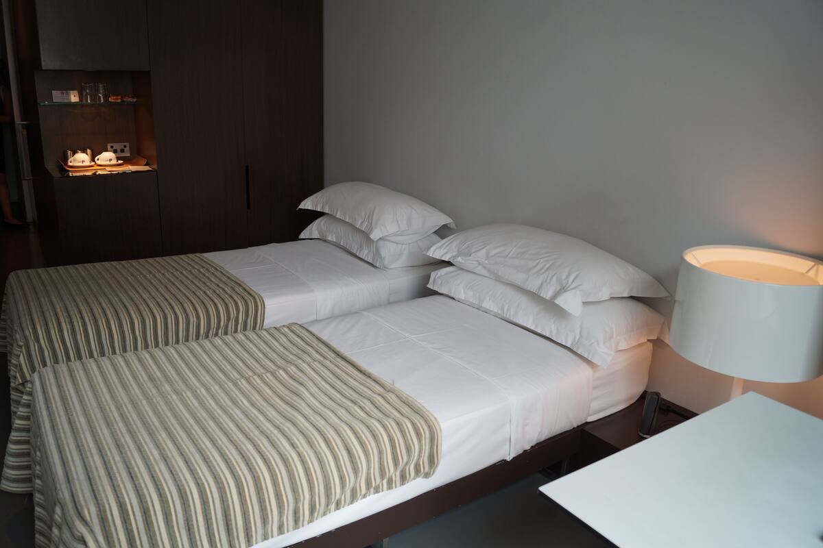 A double bed with a striped blanket, a bedside table, a lamp, and a wardrobe with a shelf.