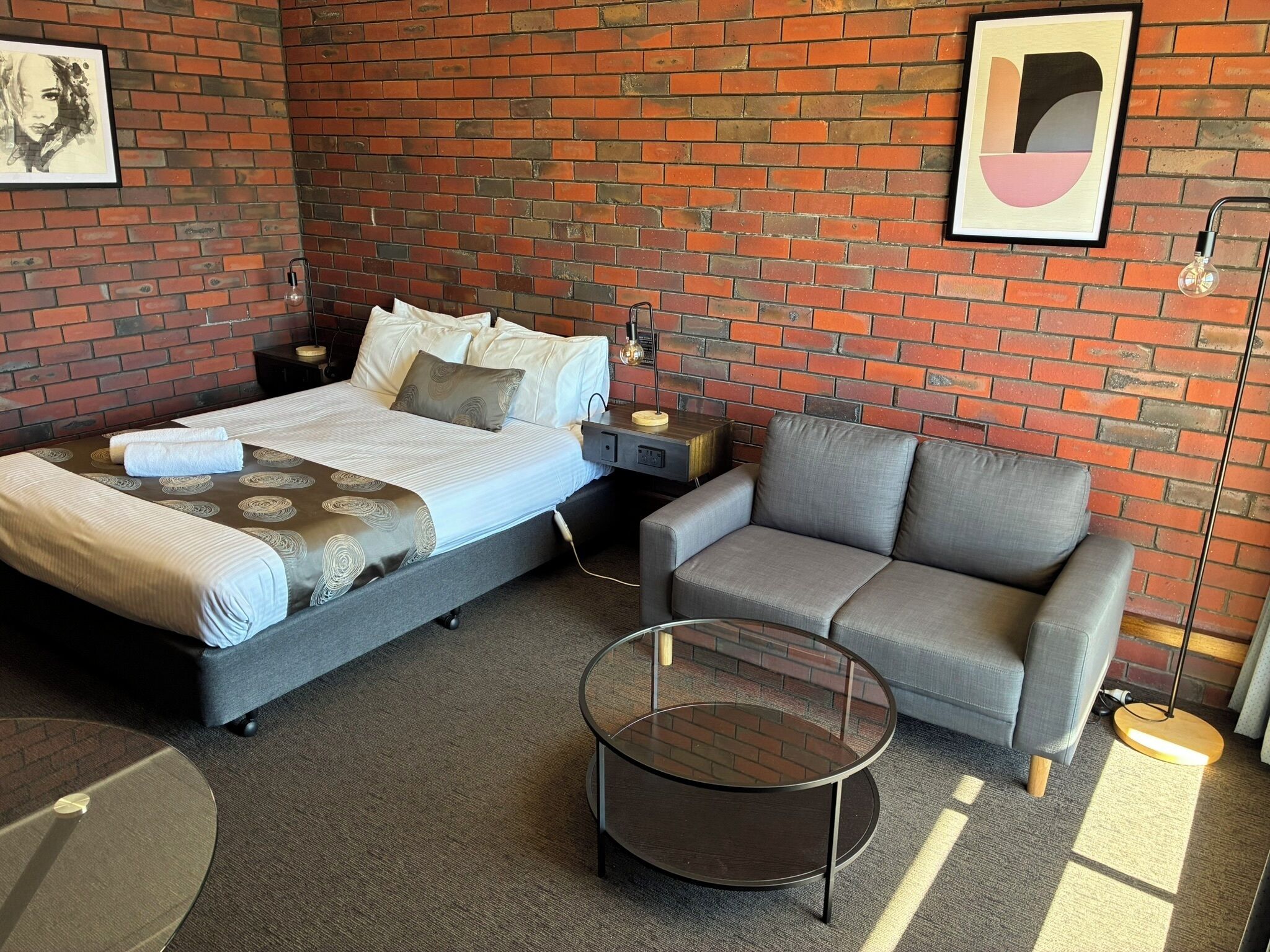 A modern hotel room with a brick wall, a bed with a grey headboard, a grey sofa, a round glass coffee table, and two framed artworks.