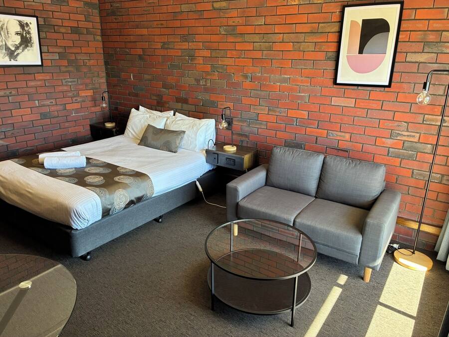 A modern hotel room with a brick wall, a bed with a grey headboard, a grey sofa, a round glass coffee table, and two framed artworks.