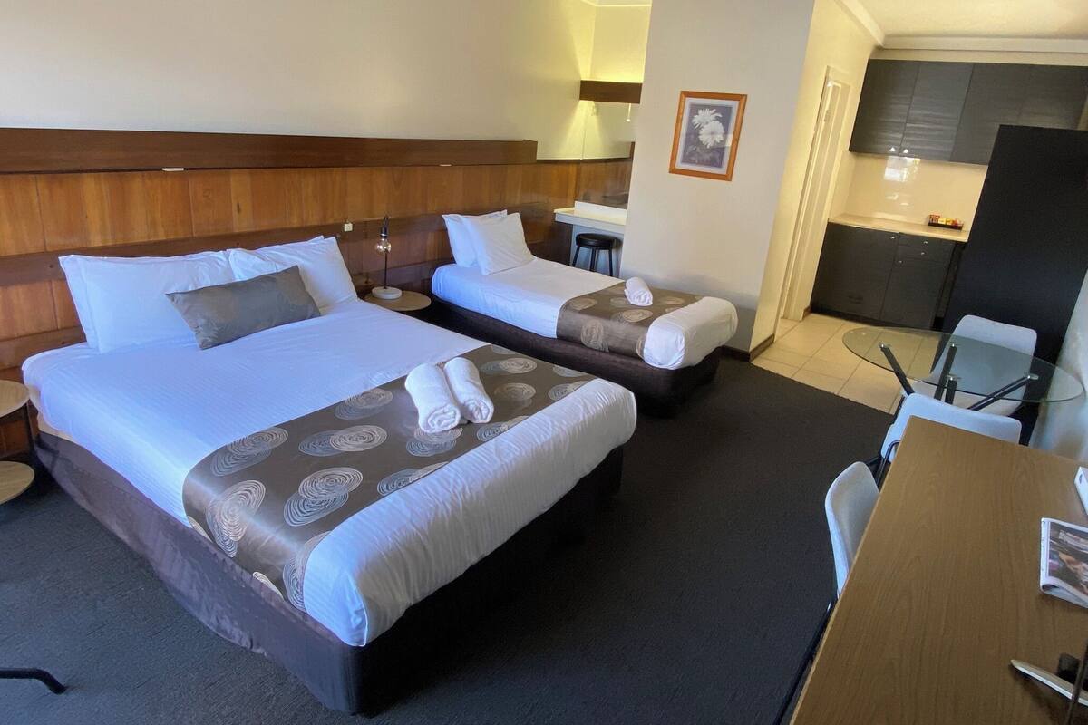A hotel room with two beds, a desk, a chair, a small table, and a kitchenette.