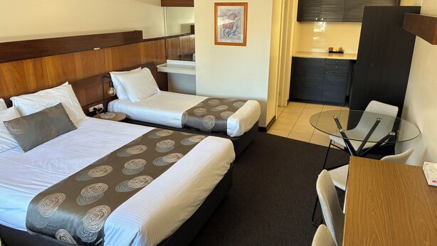 A hotel room with two beds, a small kitchenette, a glass table, and a chair. - Hi Way Eight Motor Inn (Stawell)