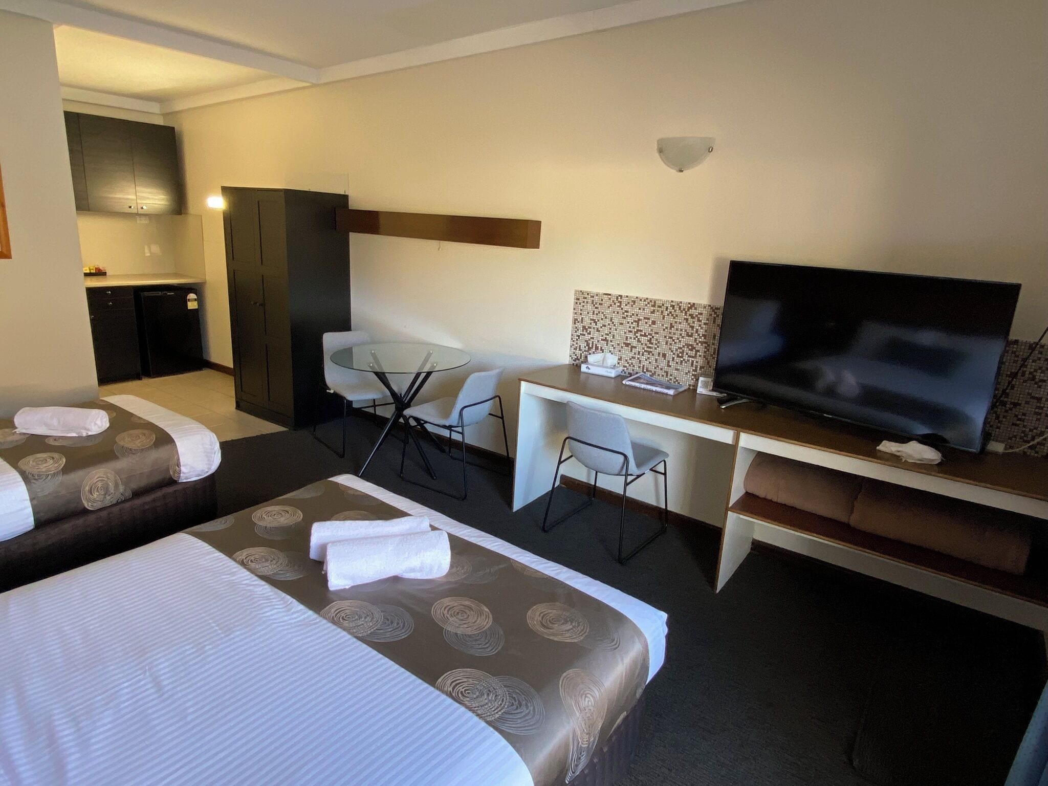 Ett hotellrum med två sängar, ett skrivbord med en TV, ett litet bord och ett litet kök.