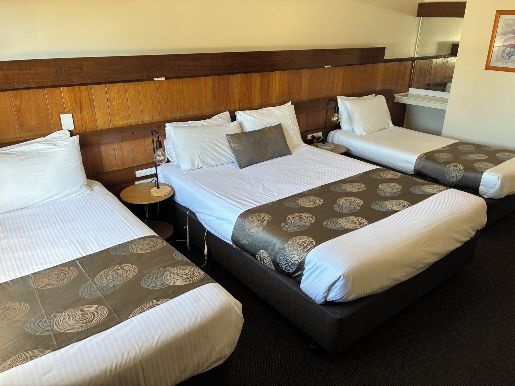 A hotel room with three beds, each with a headboard, bedside tables, and a lamp.