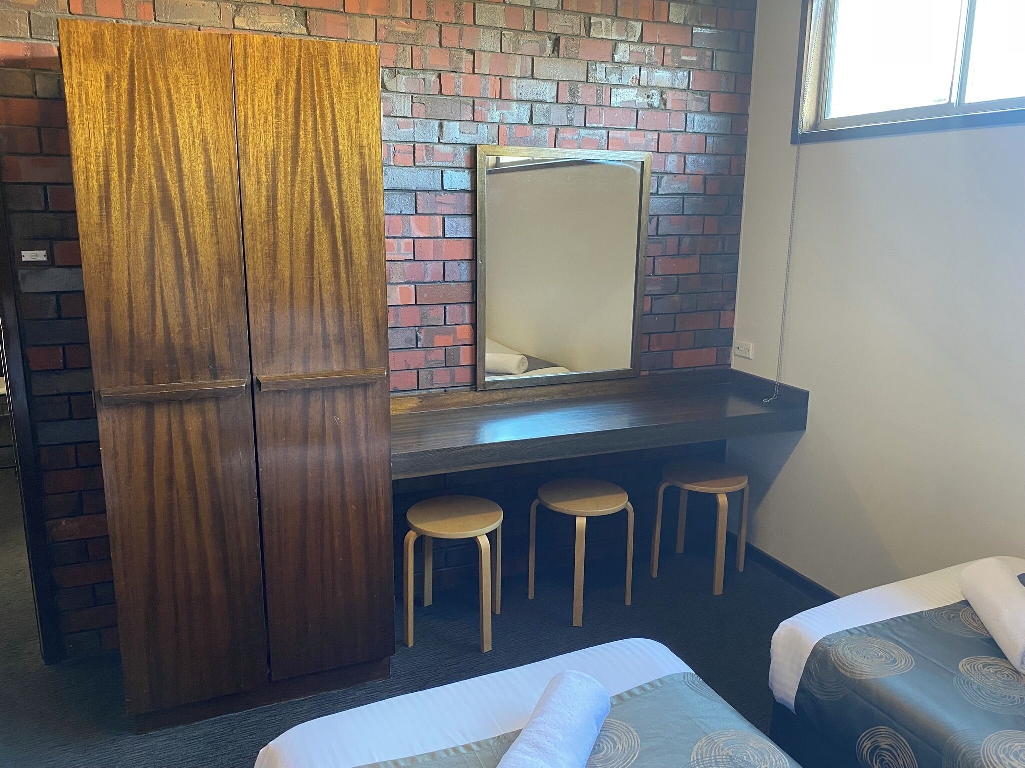 Une chambre d’hôtel avec un mur en briques, une armoire en bois, un bureau avec des tabourets et deux lits.