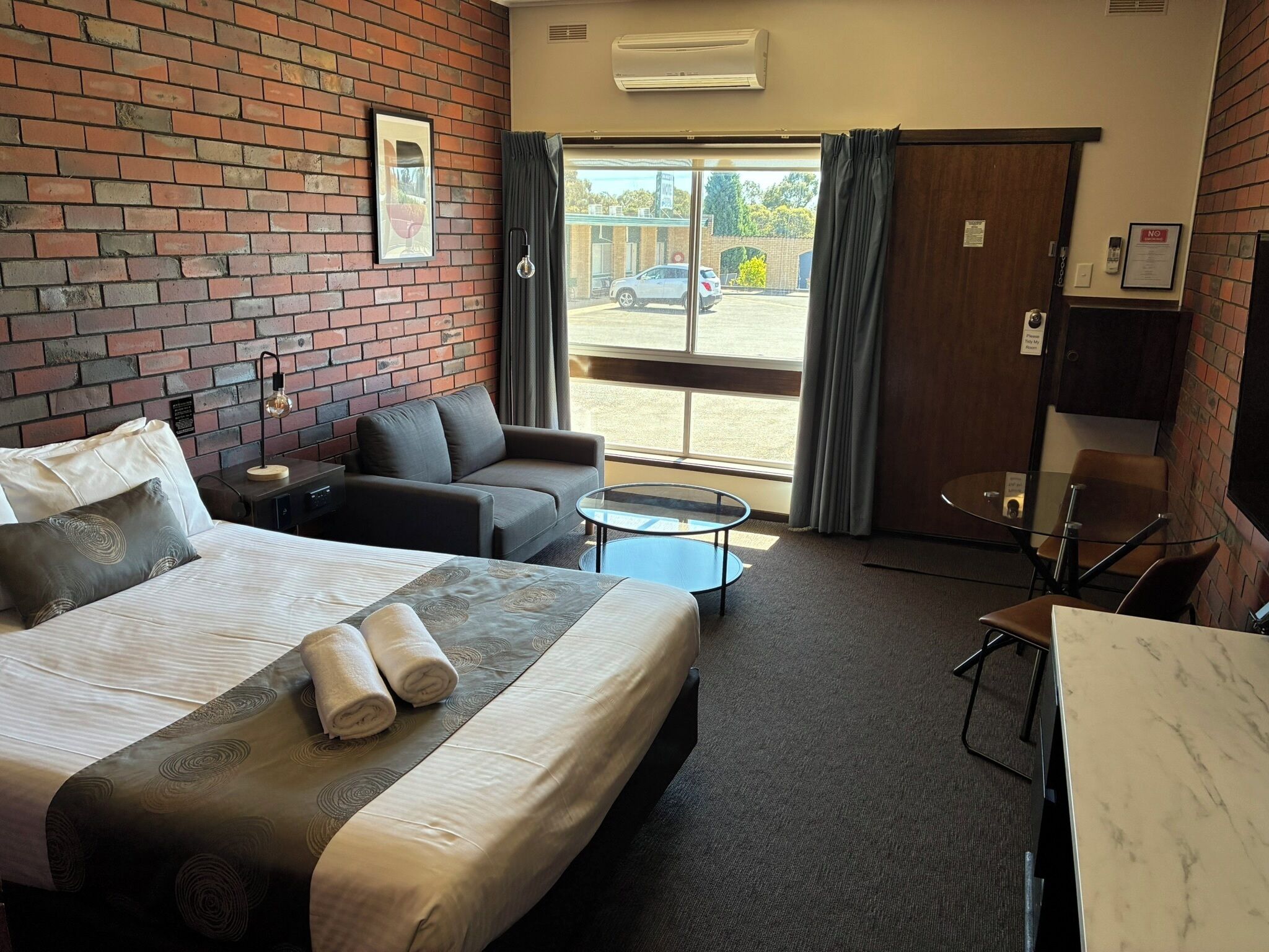 Habitación de hotel con cama, sofá, una pequeña mesa y un escritorio. Se ve una ventana con cortinas y vistas al exterior.