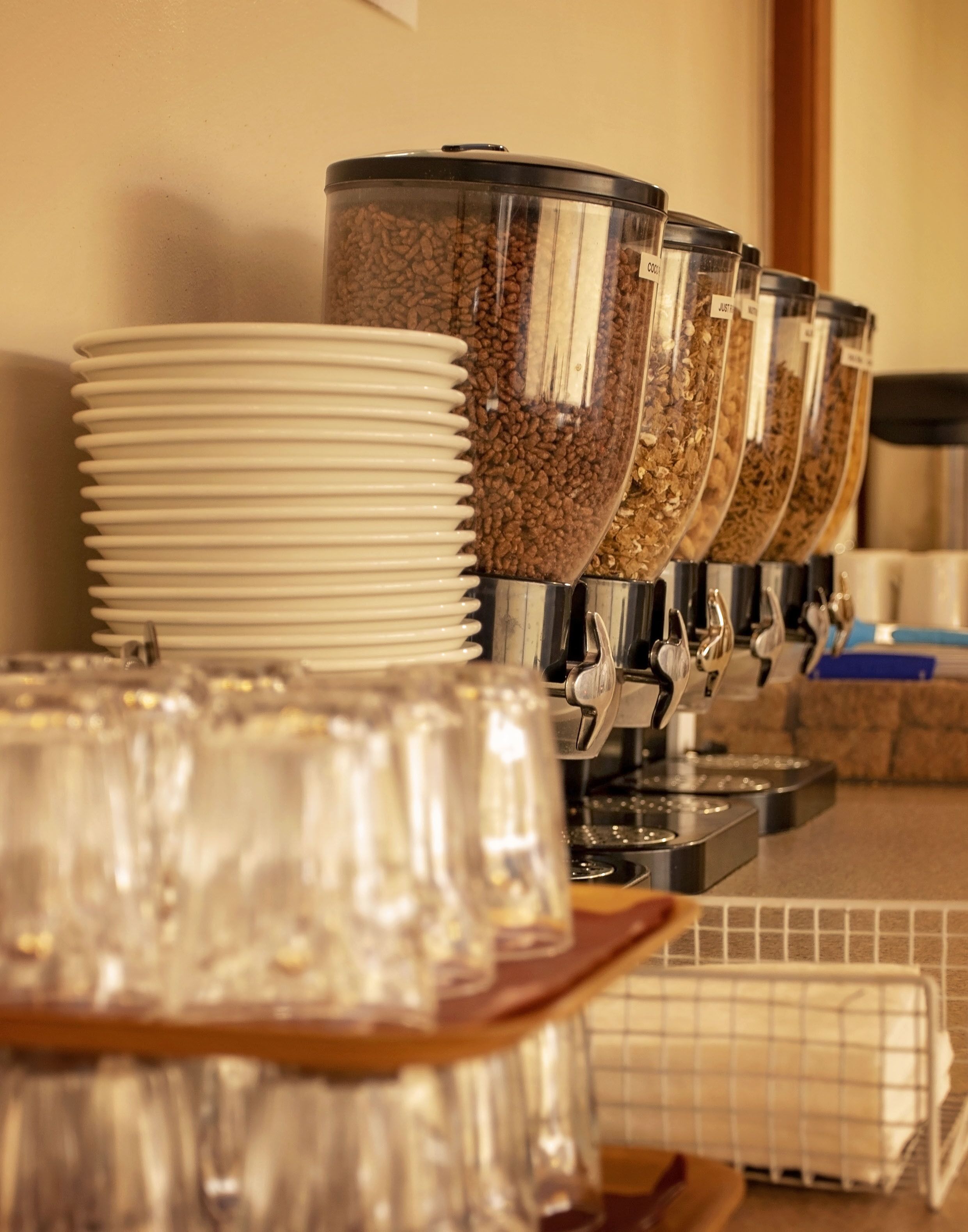 A coffee station with multiple dispensers filled with coffee beans and grounds, surrounded by stacked white plates and clear glasses.
