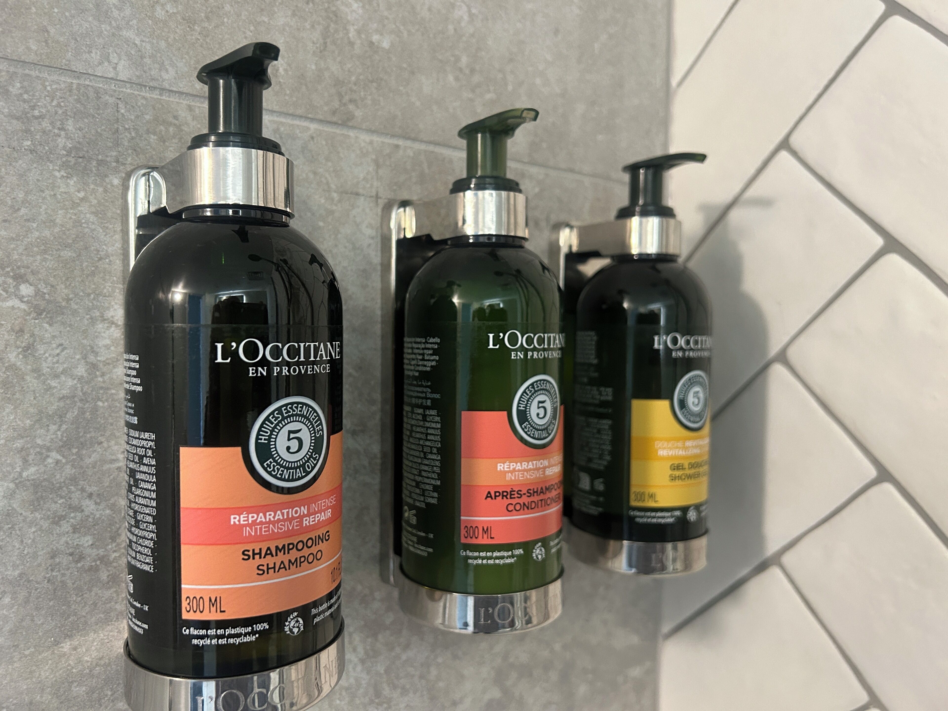 Tres botellas de champú L'OCCITANE sobre una pared de azulejos.