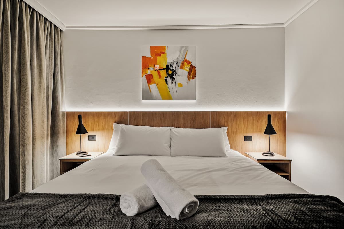 A hotel room with a large bed, two bedside tables with lamps, a painting on the wall, and a rolled towel on the bed.