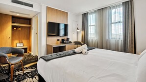 Habitación de hotel con cama, escritorio con televisor de pantalla plana, silla, armario y ventana grande con cortinas.