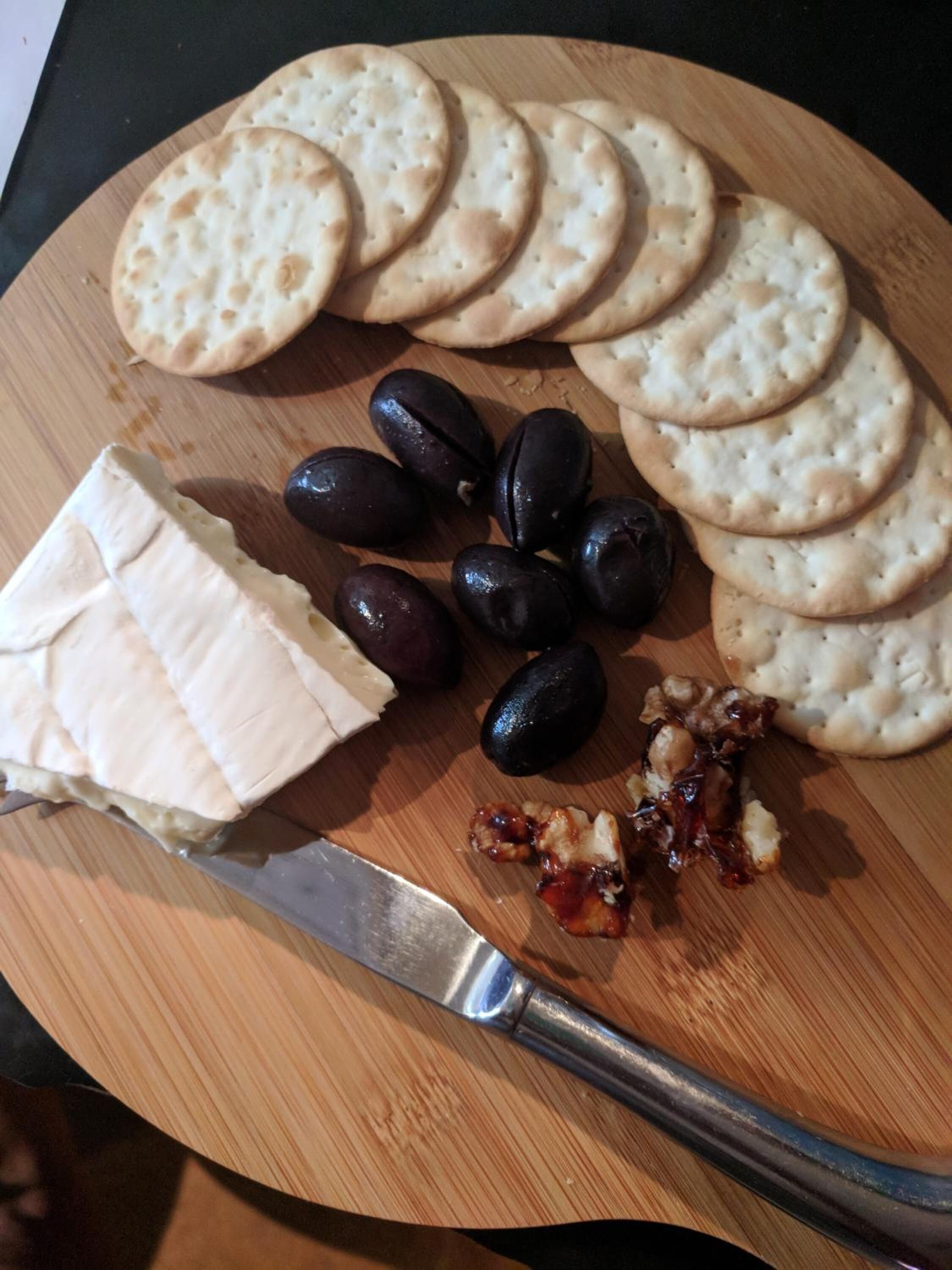 Una tabla de madera con queso, aceitunas, galletas y un cuchillo.