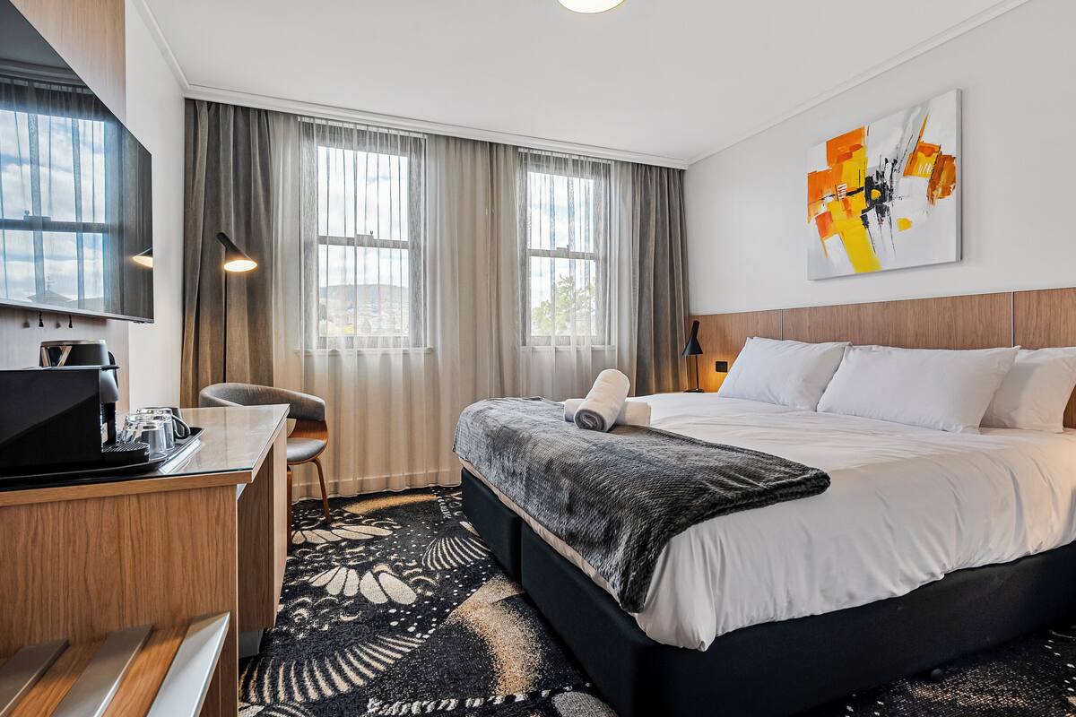 A hotel room with a bed, a desk with a computer, a chair, and a large window with curtains.
