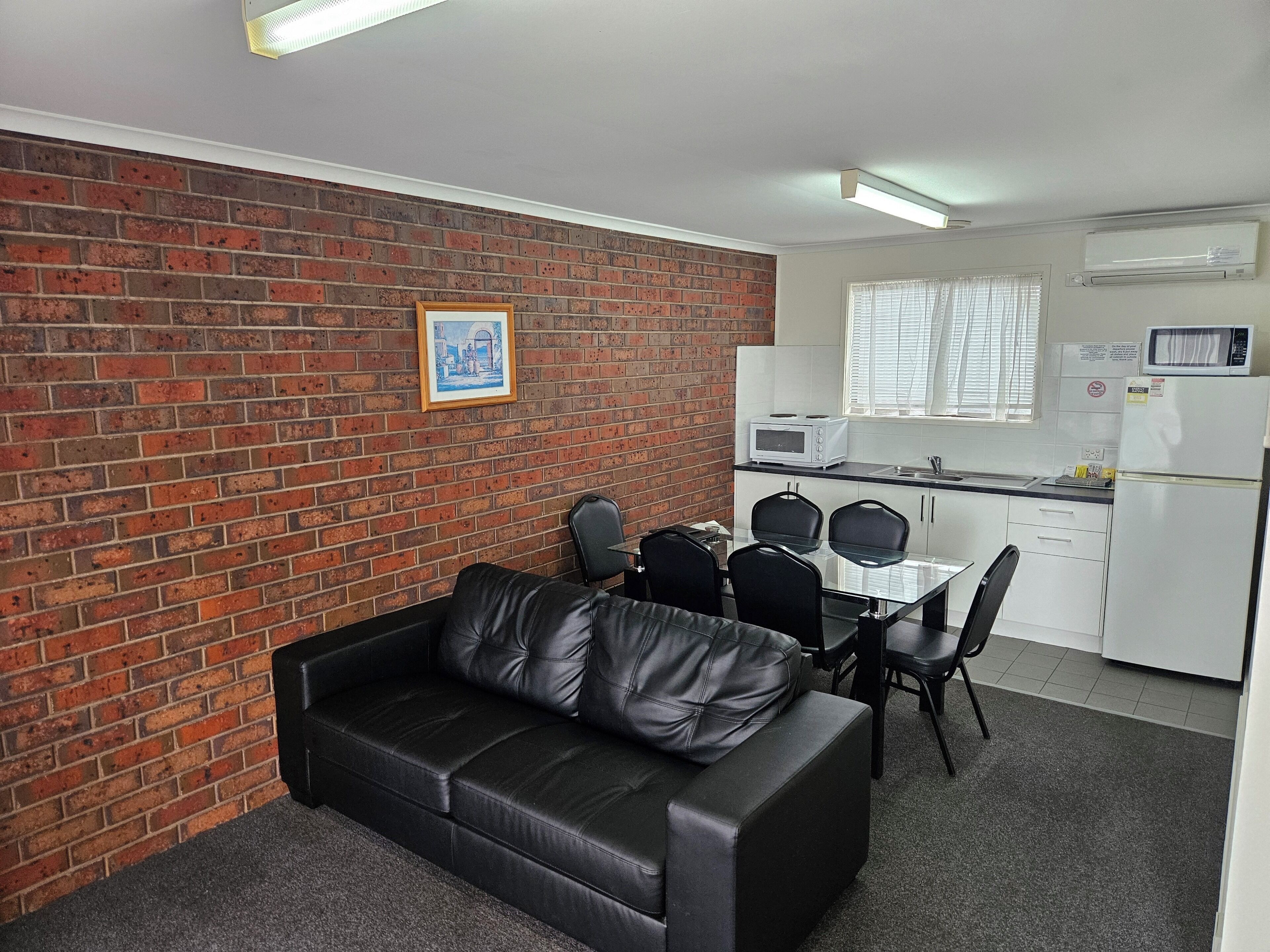 A compact living space with a brick wall, a black leather sofa, a dining table with chairs, a kitchenette with a microwave and refrigerator, and a framed picture on the wall.