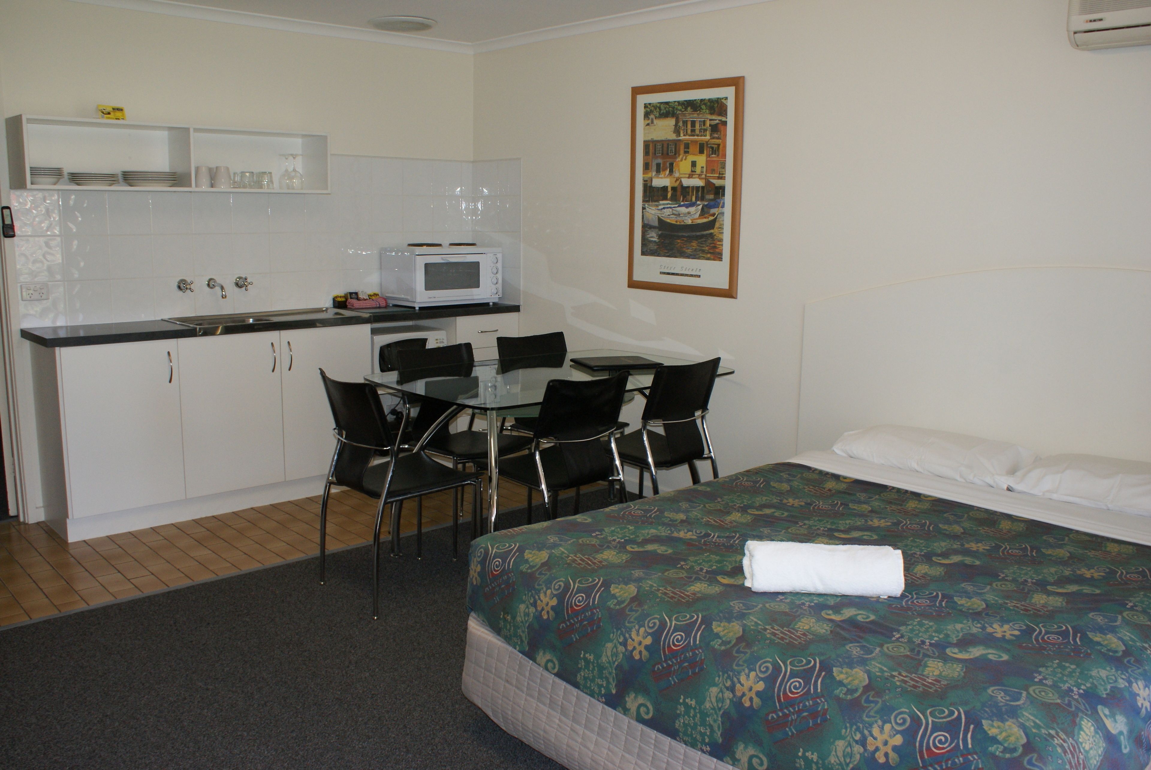 Quarto de hotel com cama, escrivaninha com cadeiras, micro-ondas e pequena cozinha com armários e pia.