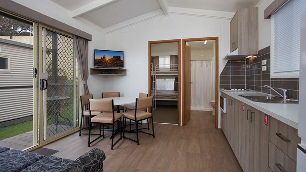 A compact kitchen with a dining area, a TV mounted on the wall, and a sliding glass door leading to an outdoor area. - Warrnambool Motel and Holiday Park (Warrnambool)