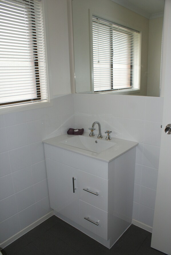 Un baño con un lavabo blanco, dos cajones, un espejo sobre el lavabo y una ventana con persianas.