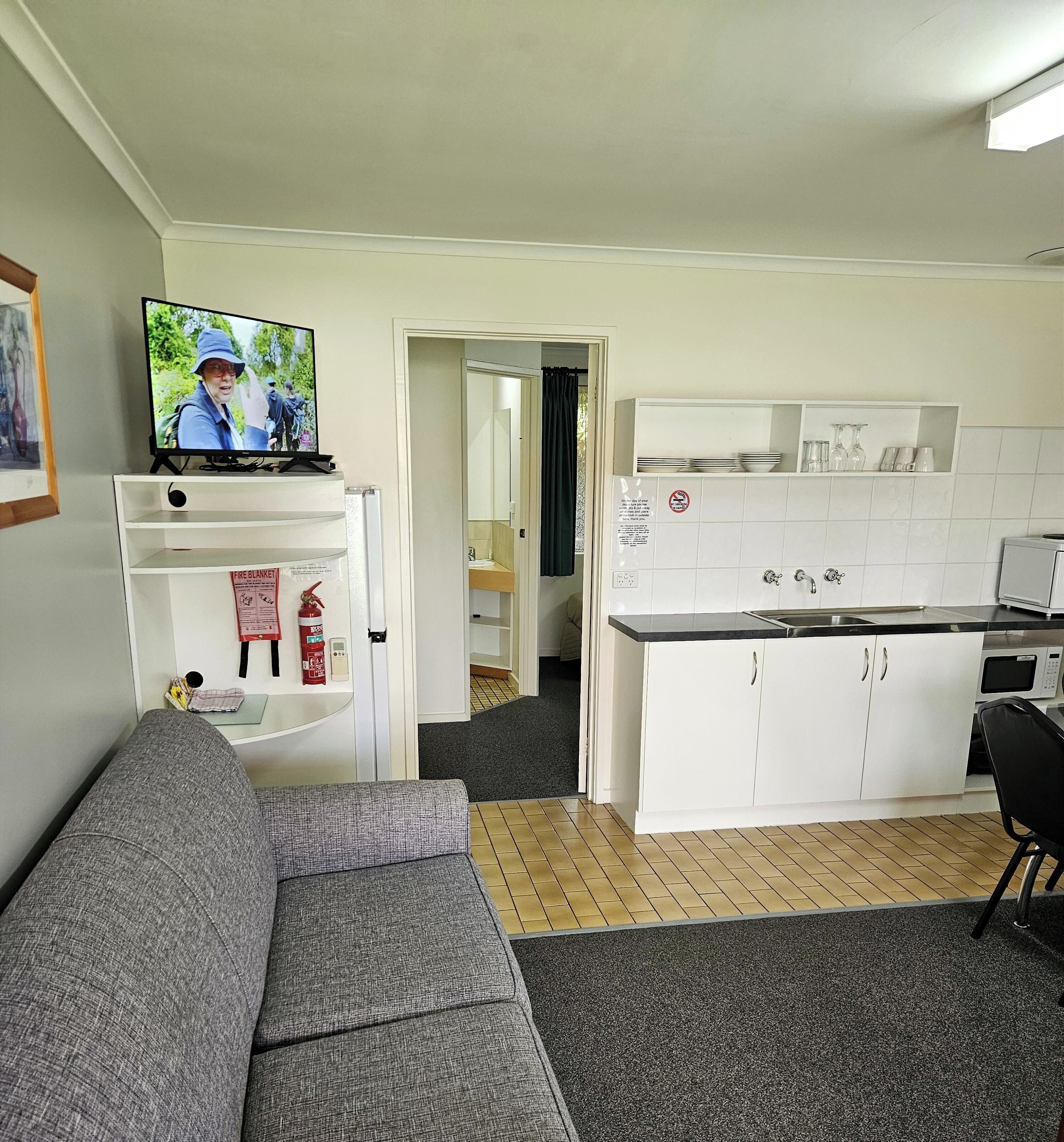 A compact living space with a grey sofa, a kitchen area with white cabinets and a black countertop, and a television on a shelf.