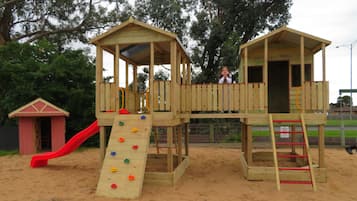 Une structure de jeu en bois, dotée d’un toboggan et d’un mur d’escalade, entourée de sable.