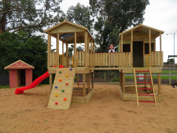 A wooden play structure with a slide and climbing wall, surrounded by sand. - Warrnambool Motel and Holiday Park (Warrnambool)
