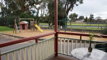 Un parque infantil con tobogán y columpios, una zona vallada y una mesa con planta en un balcón.