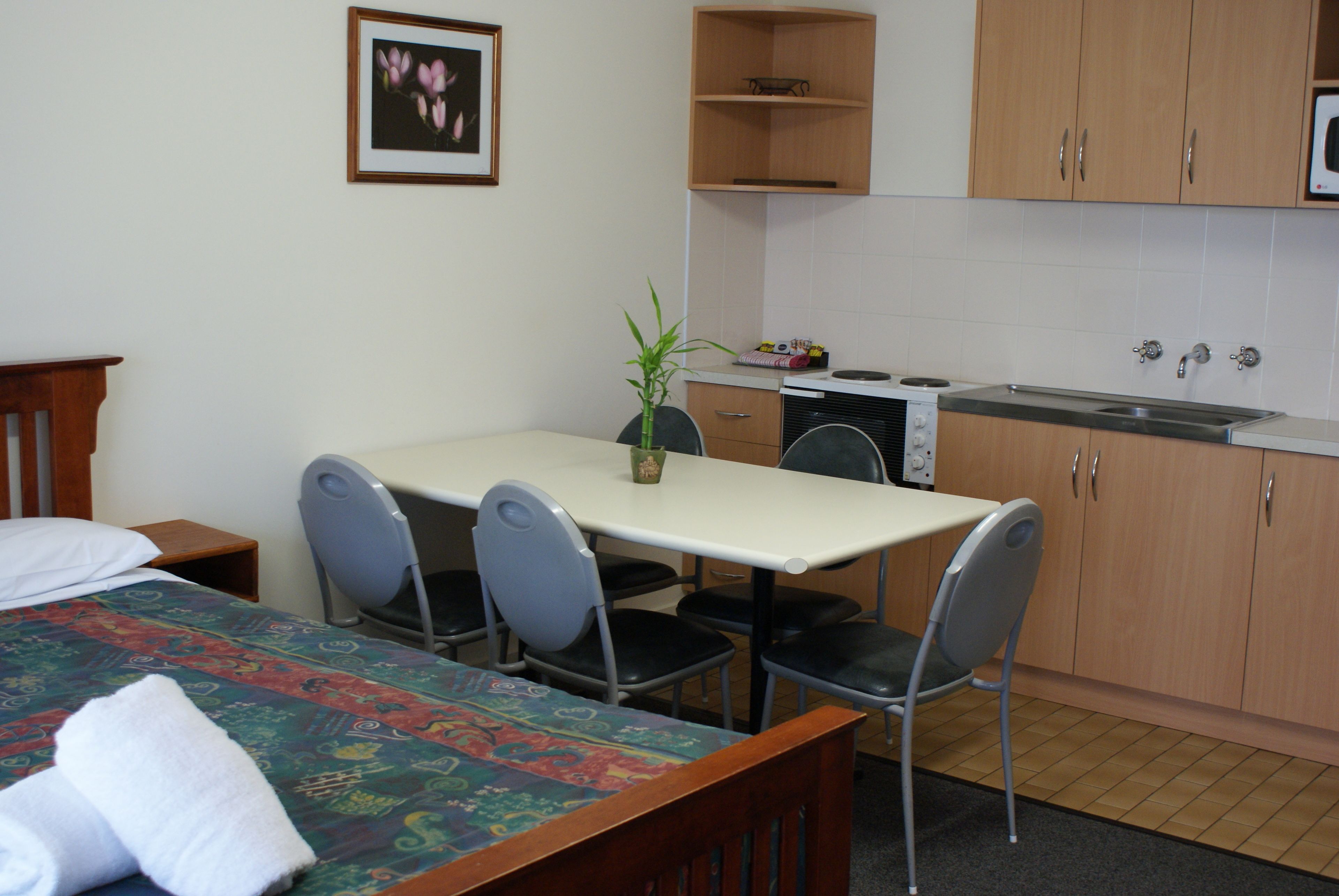 A hotel room with a bed, a dining table with chairs, a small kitchen area with a stove and sink, and a potted plant.
