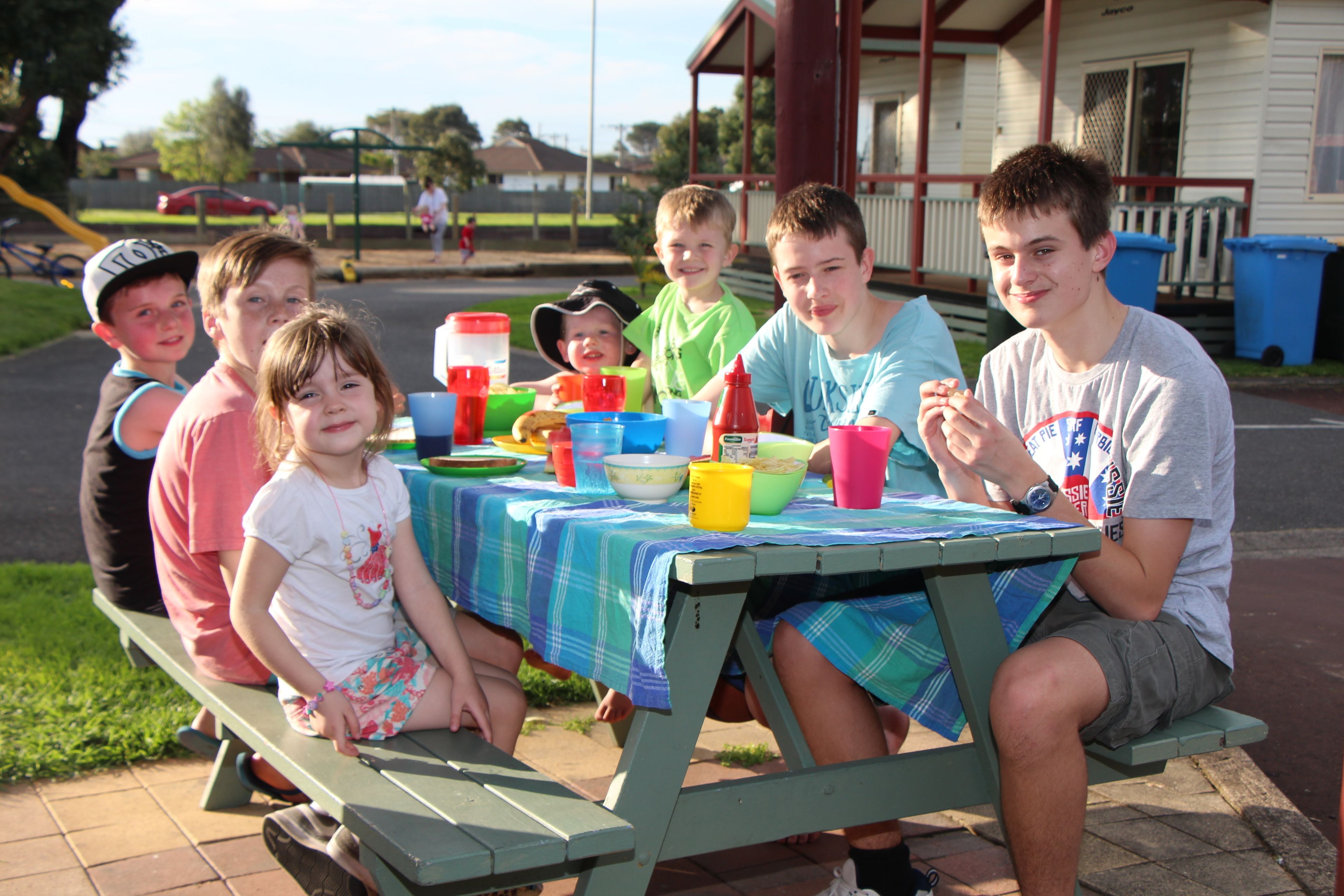 A group of people sitting at a picnic table with various food items and drinks.