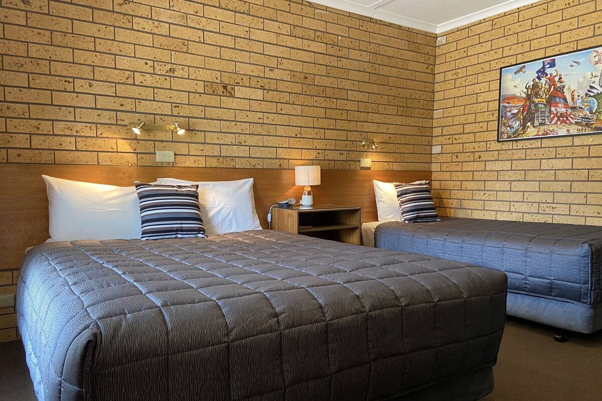 A hotel room with two beds, a brick wall, a painting, and a nightstand with a lamp.