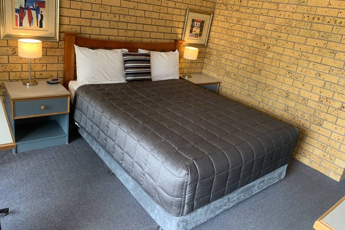 A hotel room with a brick-patterned wall, a bed with a dark quilted bedspread, two bedside lamps, and a small nightstand with a clock.