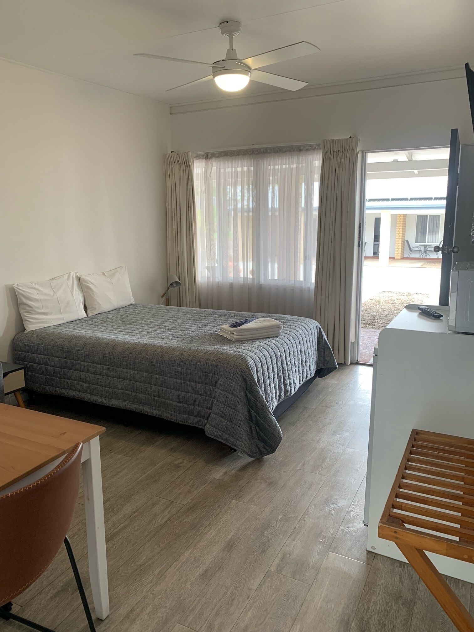 A hotel room with a bed, a desk, a chair, a ceiling fan, and a view of the outside.
