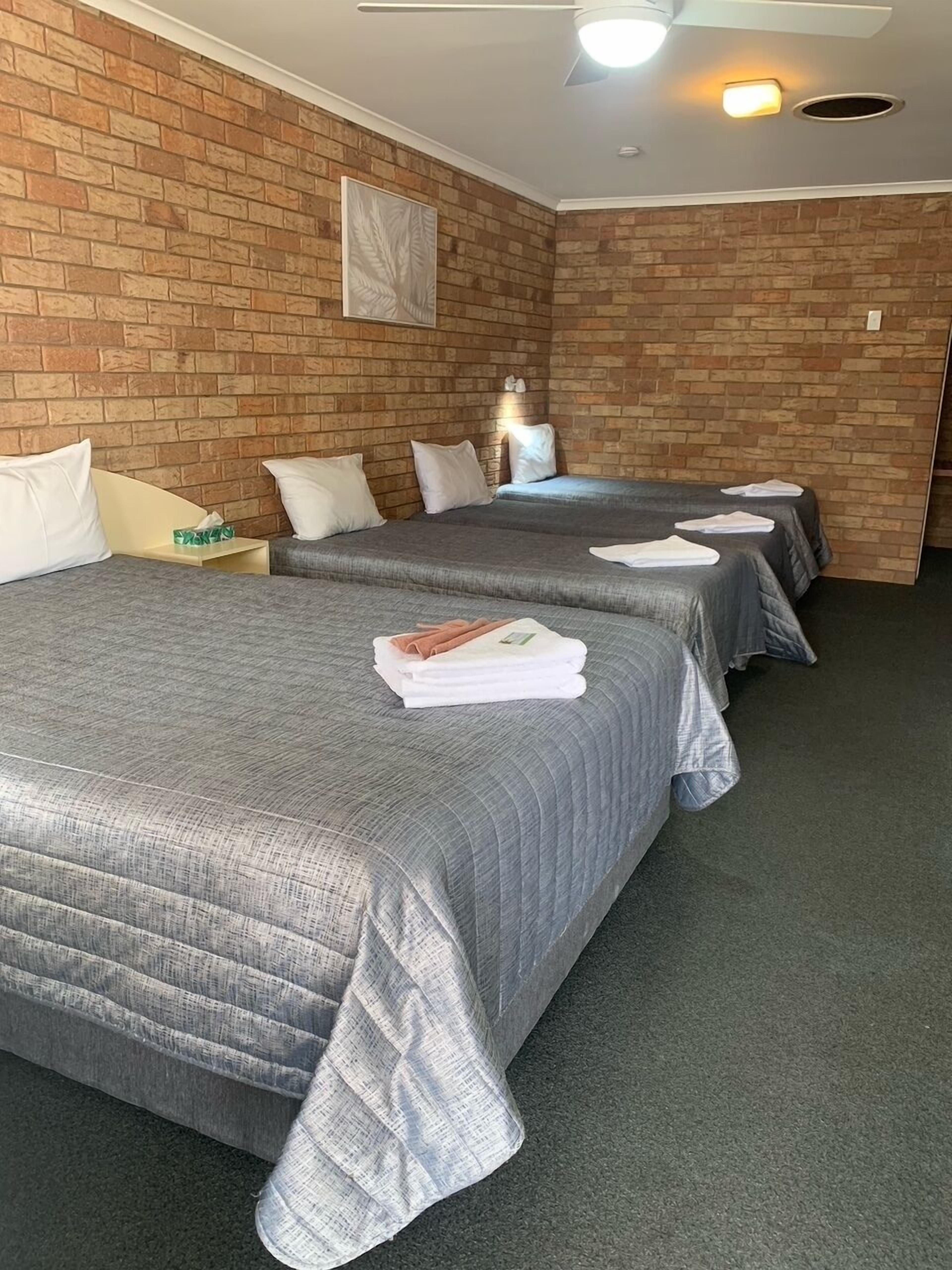 A hotel room with two beds, each with grey bedding and white pillows. The room has brick walls and a ceiling fan.