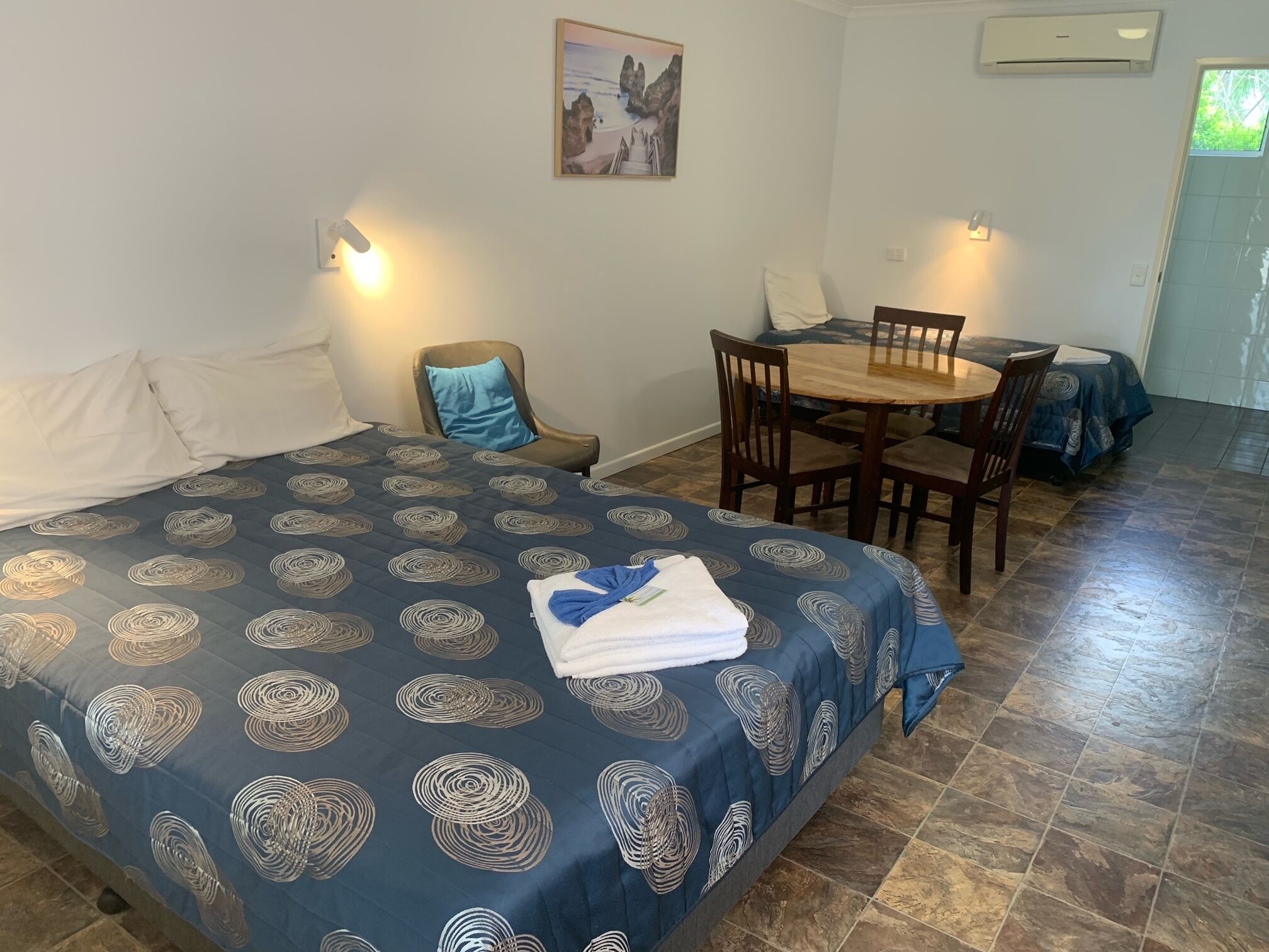 Quarto de hotel com uma cama, uma mesa de jantar e cadeiras. Há um quadro na parede e um ar condicionado instalado acima da mesa.