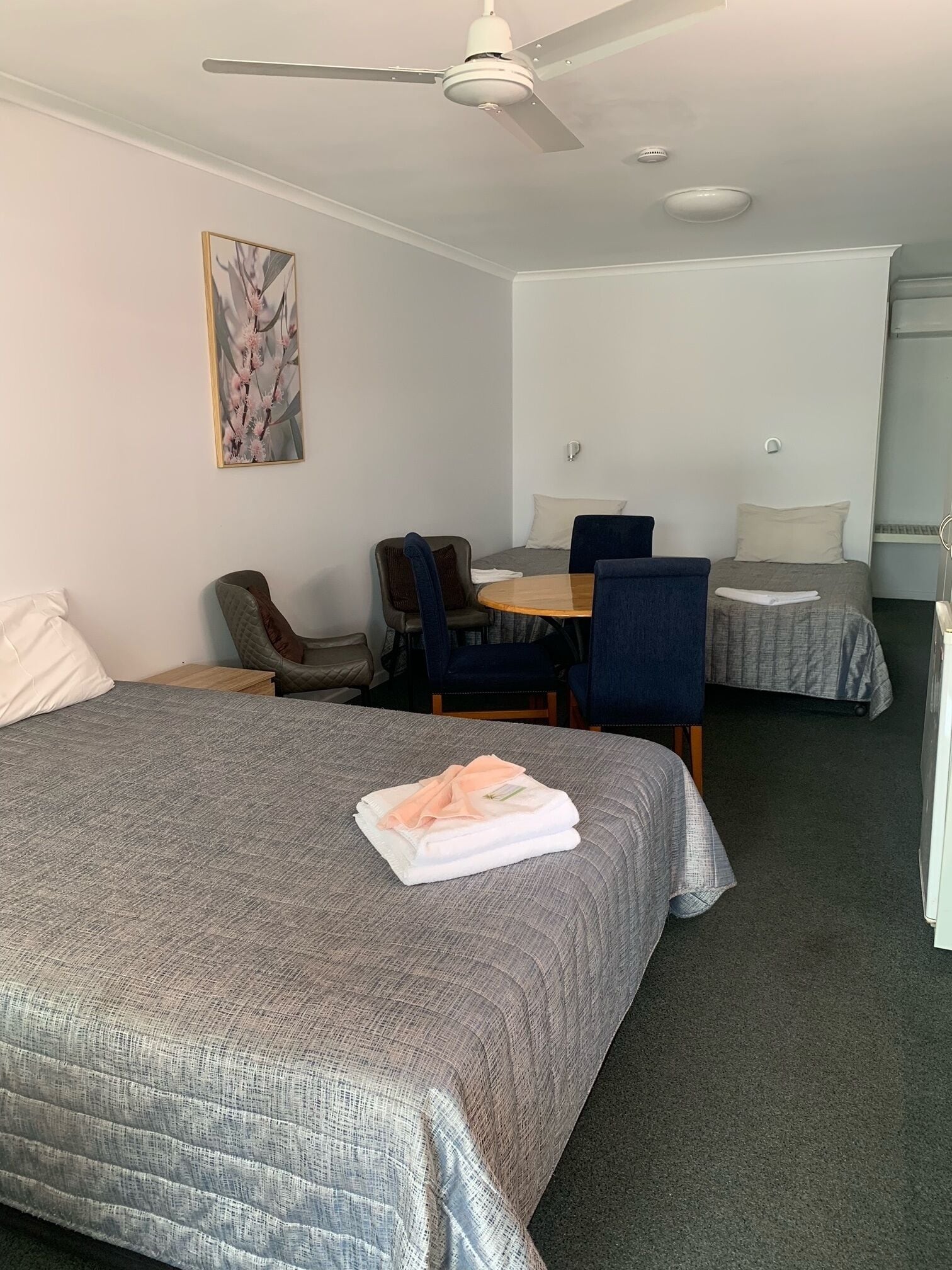 Habitación de hotel con una cama grande, un escritorio, dos camas más pequeñas, un ventilador de techo y una obra de arte colgada en la pared.