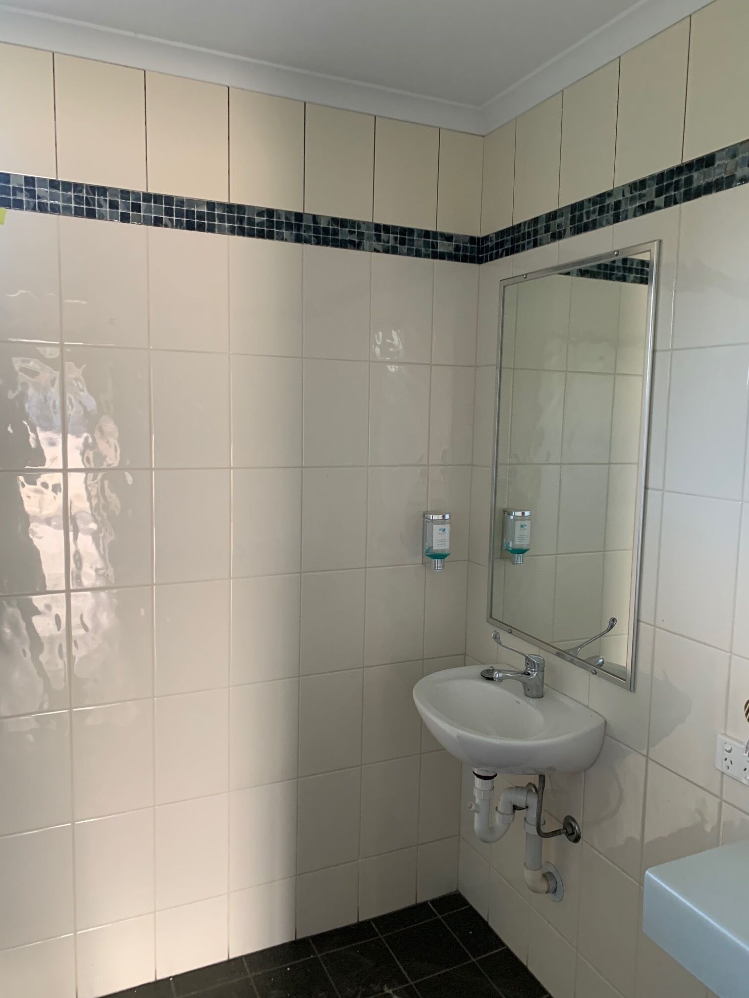 Une salle de bain avec des carreaux de céramique blancs, un sol carrelé de noir, un lavabo blanc, un miroir et deux distributeurs de savon.