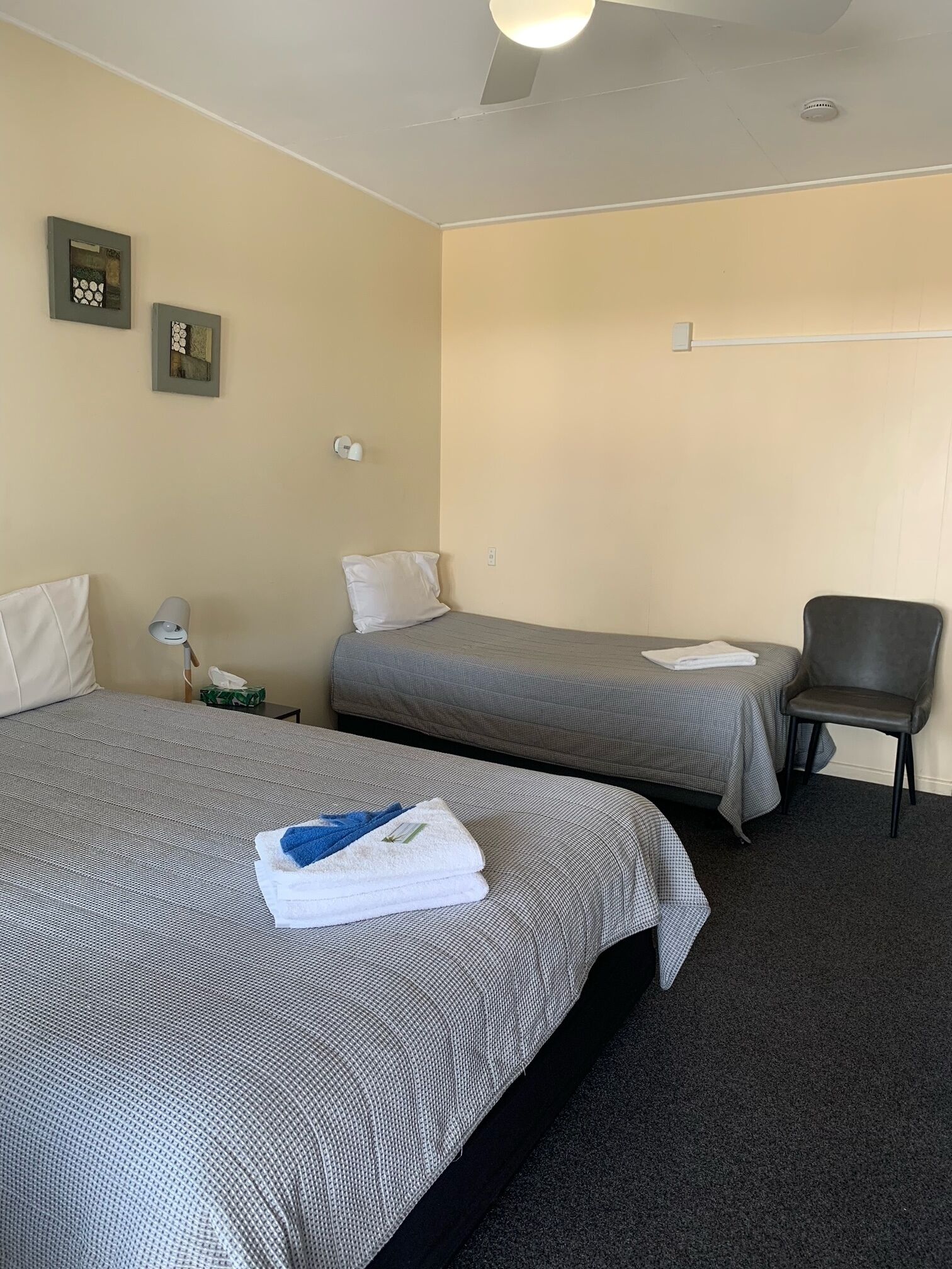A hotel room with two single beds, a chair, a nightstand, and a wall-mounted light fixture.