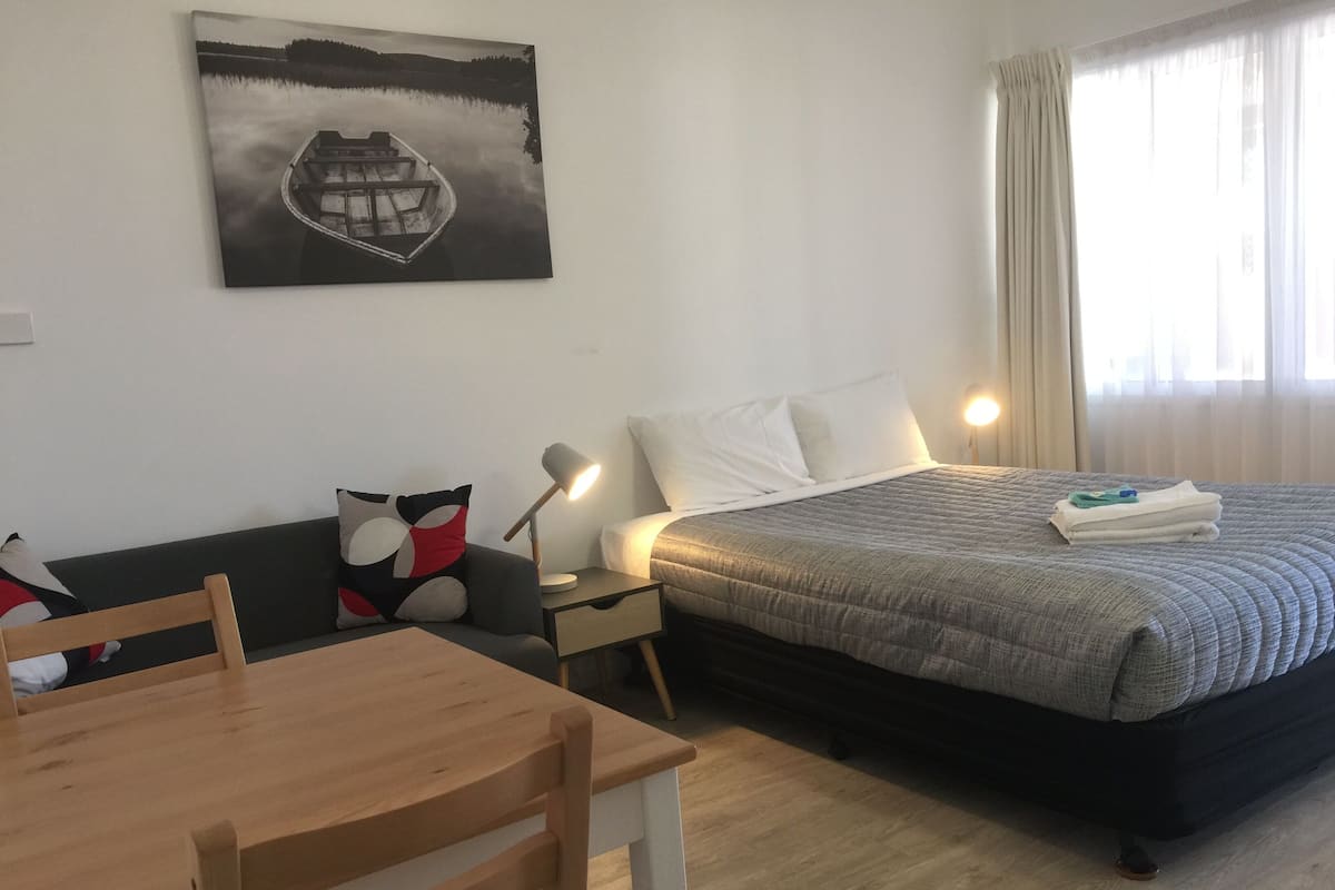 Una habitació d'hotel amb un llit, escriptori, cadira, taulell de nit amb una làmpada, un quadre a la paret i una finestra amb cortines.