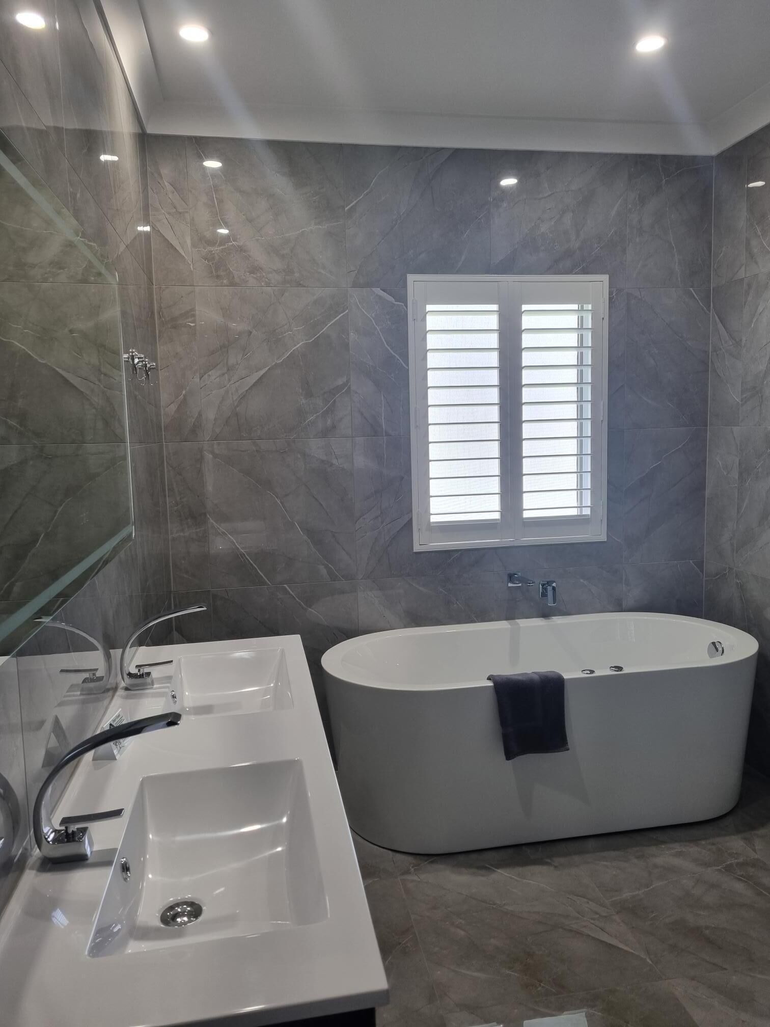 A modern bathroom with a freestanding bathtub, a large mirror, and a window with blinds.