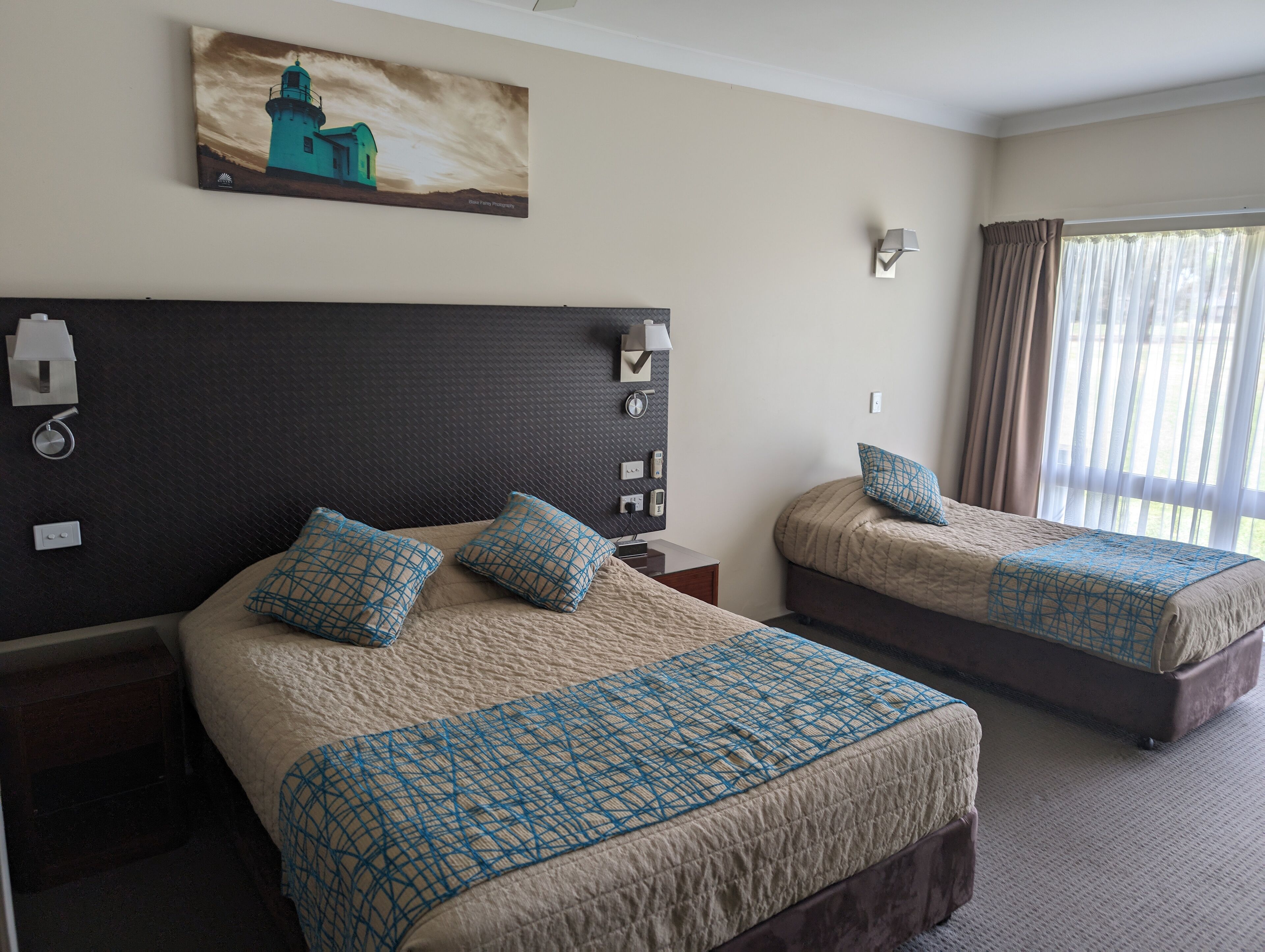 Une chambre d’hôtel avec deux lits, un tableau représentant un phare, une horloge murale et une fenêtre avec des rideaux.