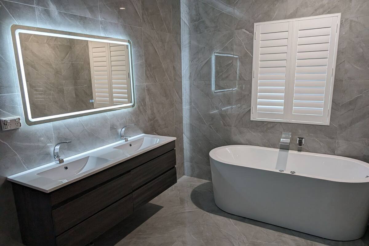 A modern bathroom with a freestanding bathtub, a vanity with a mirror, and a window with shutters.